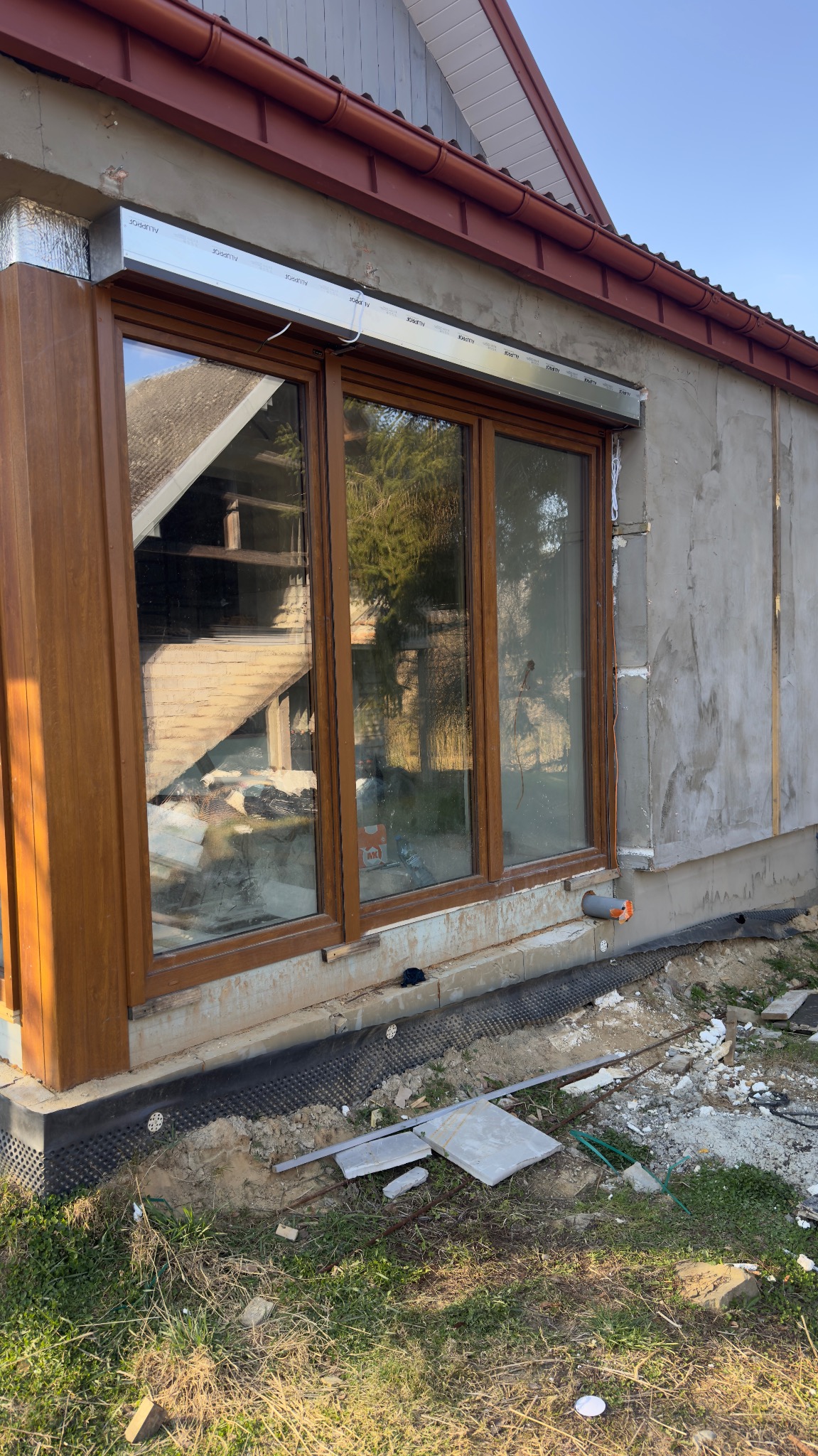 Montaż okien PCV z roletami zewnętrznymi w surowym stanie elewacji. Widoczne brązowe ramy okienne i srebrne kasety rolet. Prace wykończeniowe w toku.