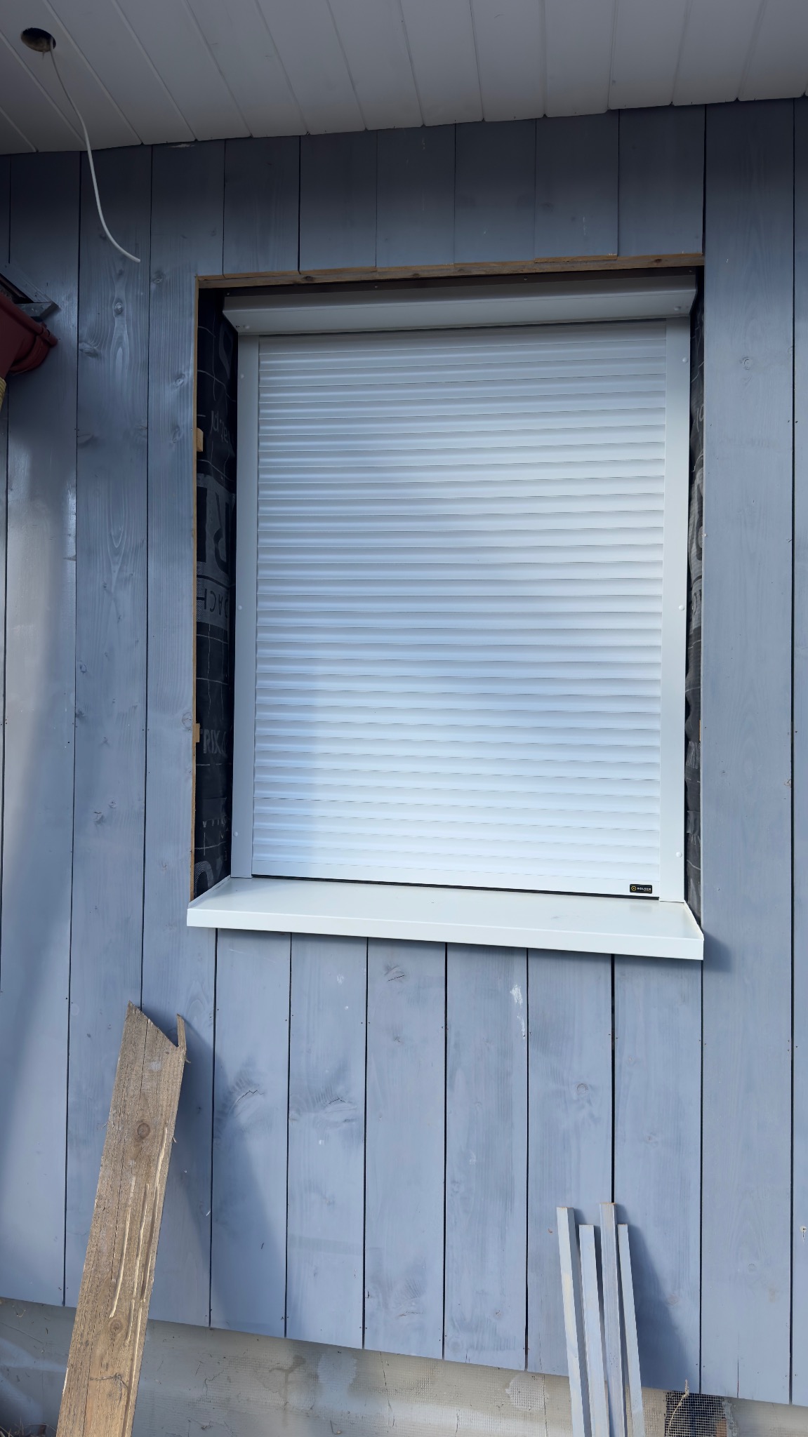 Zamknięta, biała roleta zewnętrzna na oknie w szarej, drewnianej elewacji budynku. Widoczne deski i fragmenty materiałów budowlanych.