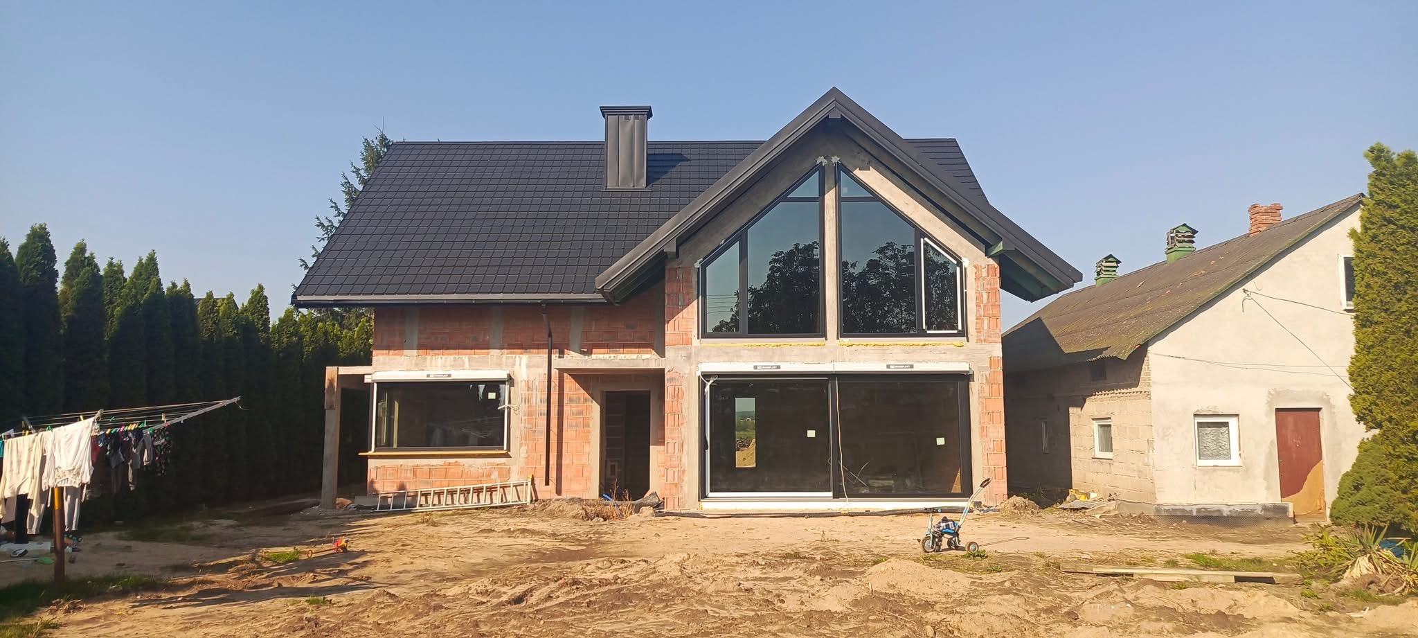 Dom w trakcie budowy z zamontowanymi oknami z czarnymi ramami i roletami zewnętrznymi. Widoczne cegły i czarny dach. Na podwórku zabawka.