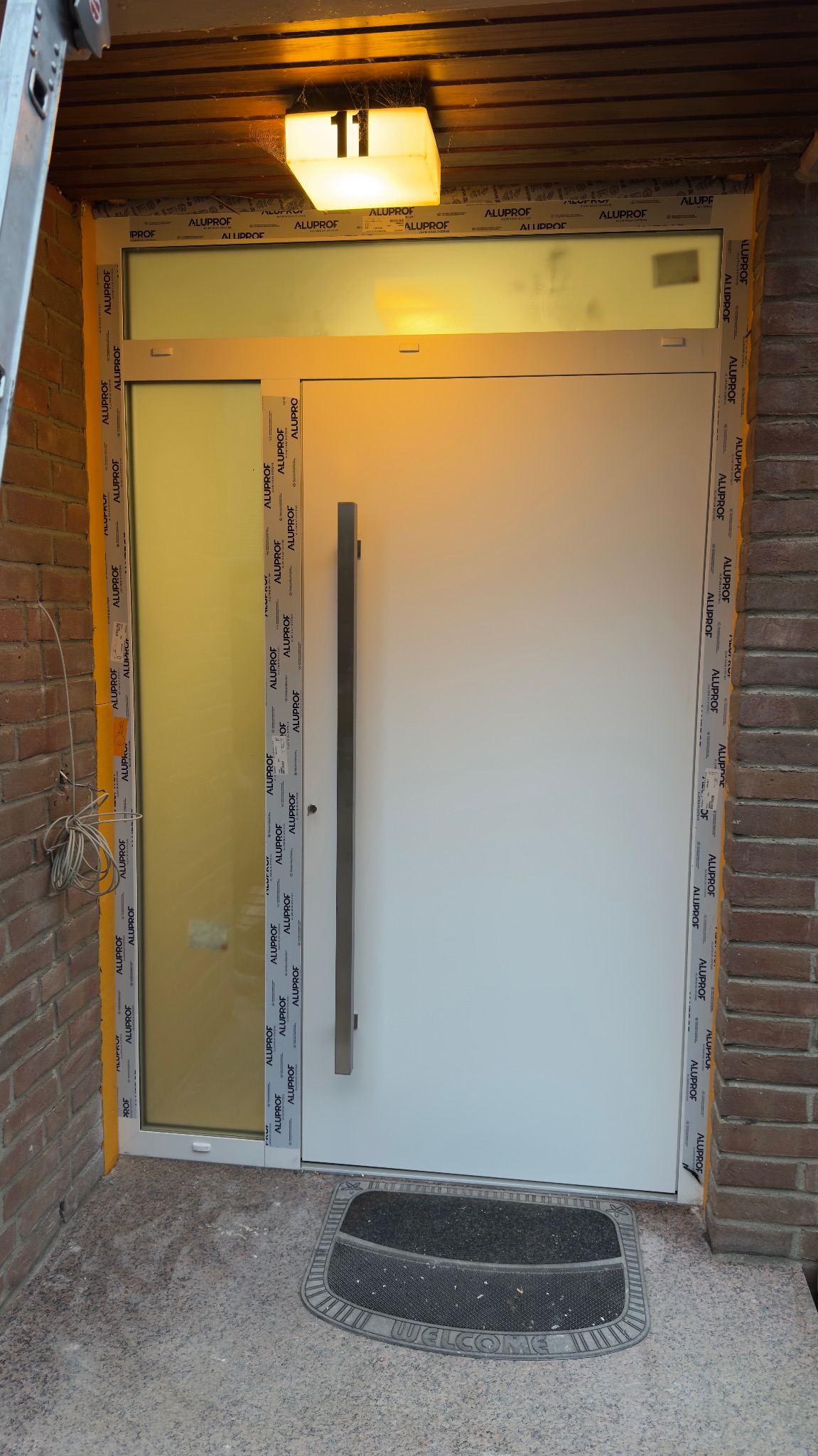 Białe drzwi zewnętrzne z długim, srebrnym uchwytem, zamontowane w ceglanym murze. Nad drzwiami lampa z numerem 11. Wykończenie ramy z logo producenta.