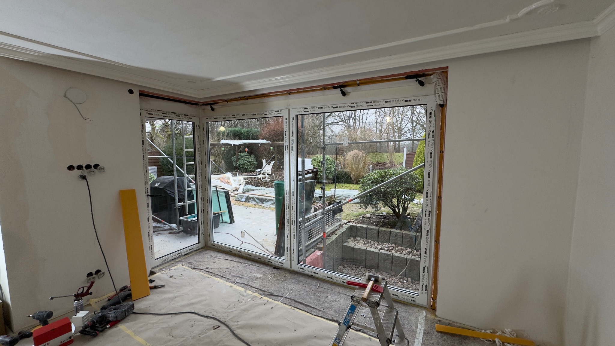 Montaż narożnych okien PCV z widokiem na ogród. Widoczne narzędzia i drabina. Etap prac wykończeniowych w jasnym pomieszczeniu z dekoracyjną sztukaterią.