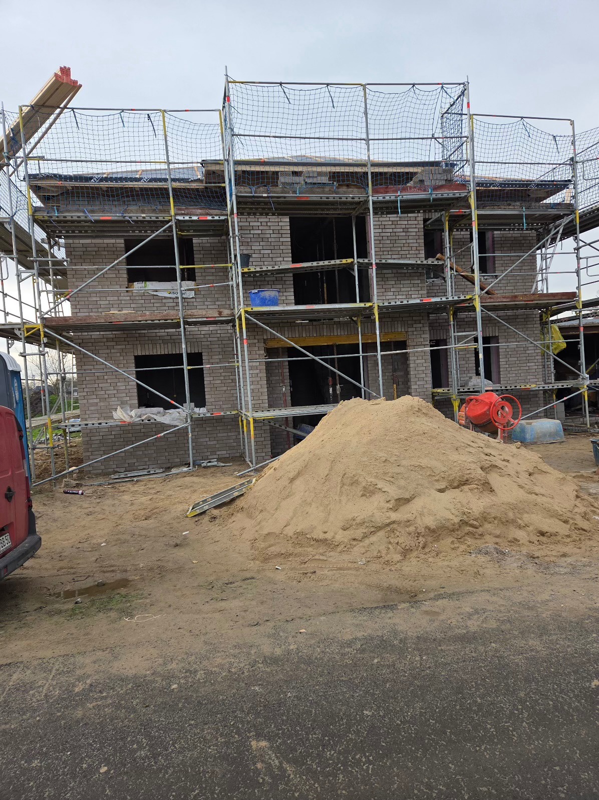 Surowy stan budowy domu z cegły, z rusztowaniami i stertą piasku na pierwszym planie. Widoczna betoniarka i częściowo zaparkowany samochód dostawczy.