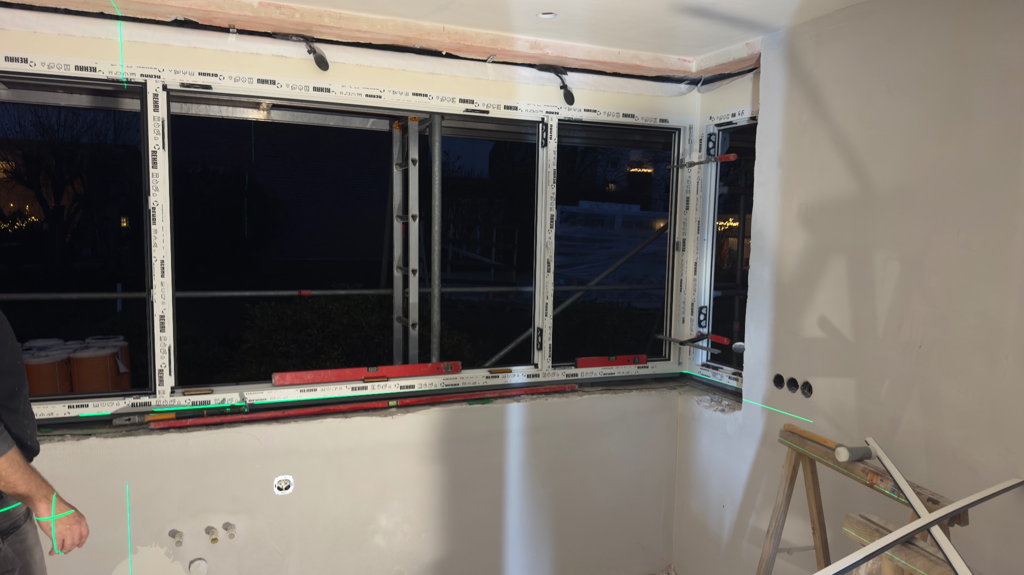 Montaż okna PCV Rehau w nowym budynku, widoczne poziomice laserowe, drabina i narzędzia. Etap prac wykończeniowych, wieczorne oświetlenie zewnętrzne.