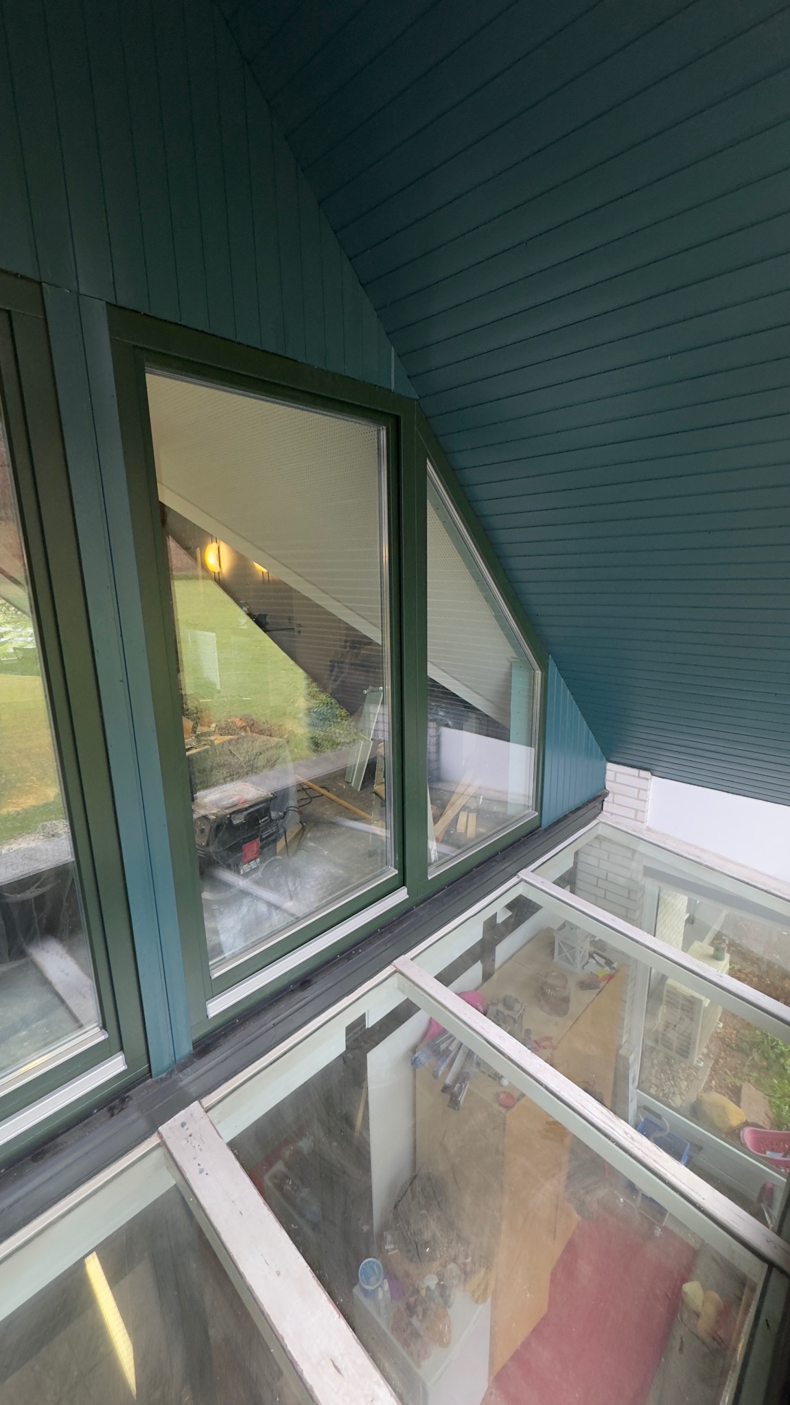 Zielone okna PCV o nietypowym kształcie w skośnej ścianie poddasza, widok z dołu na zewnątrz. Widoczne szklane panele w podłodze i elementy wyposażenia wnętrza.