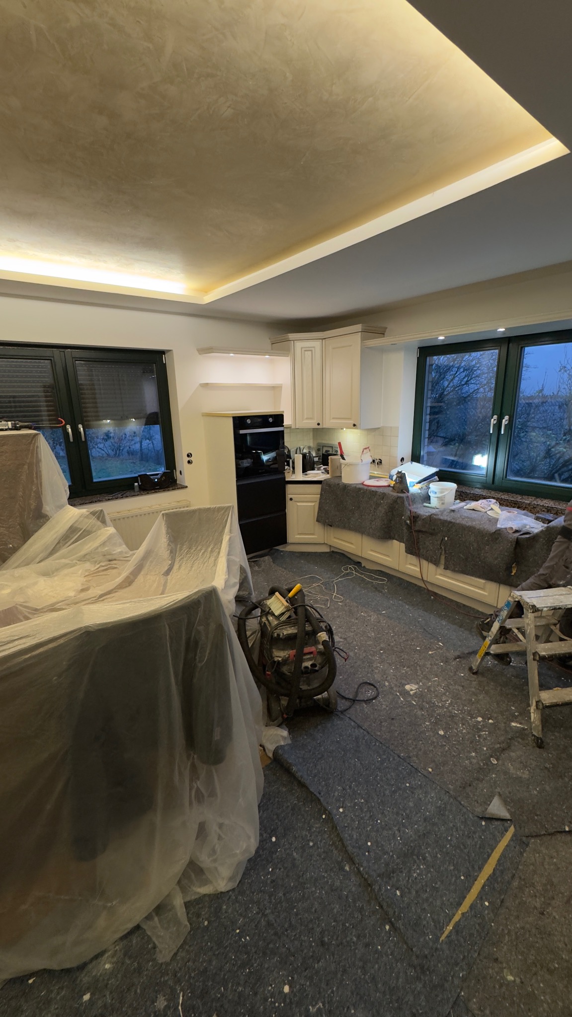 Kuchnia w trakcie remontu: meble zabezpieczone folią, widoczne narzędzia malarskie i świeżo pomalowany sufit z oświetleniem LED. Okna z widokiem na zewnątrz.