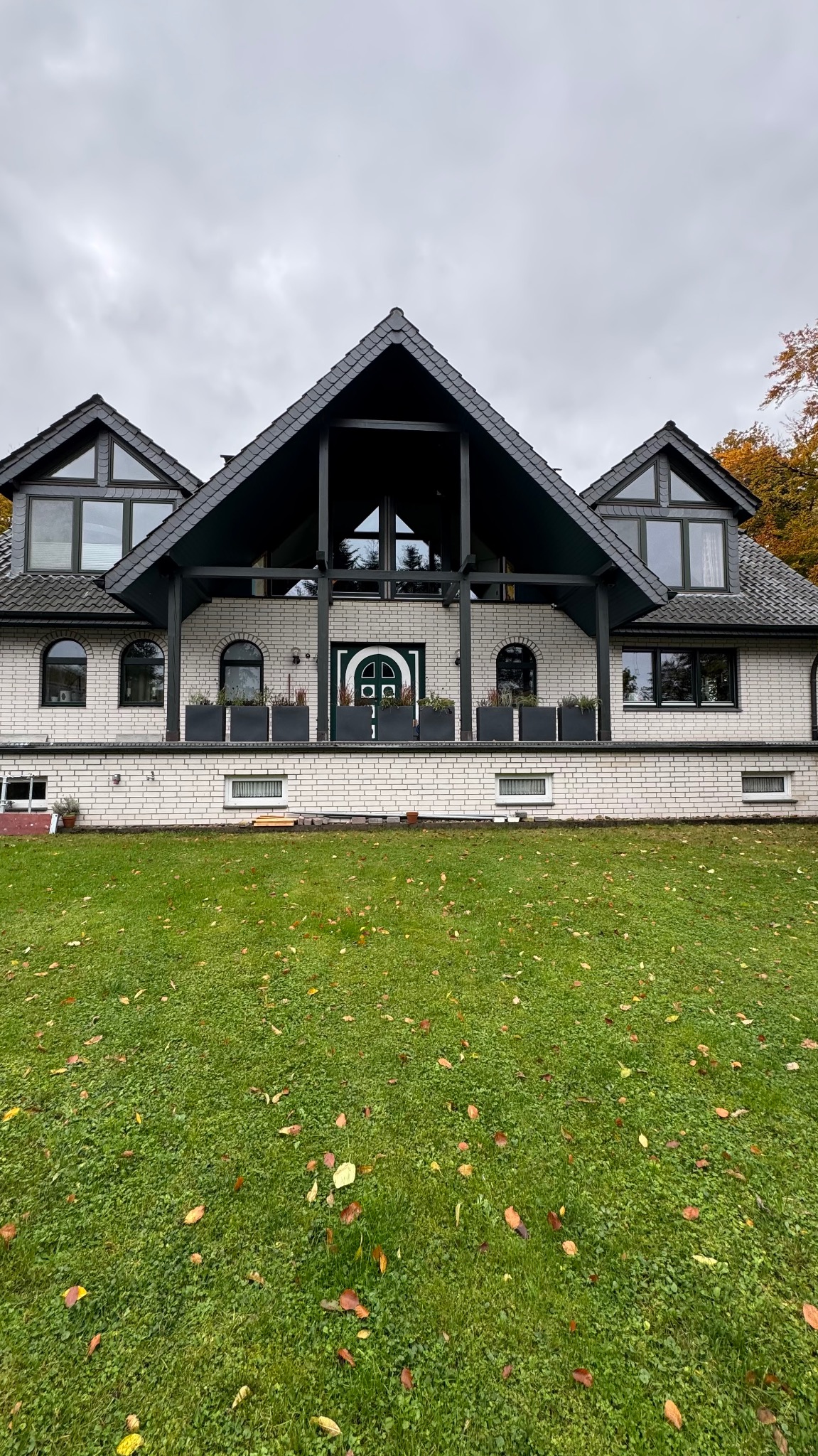 Biały dom z ciemną, spadzistą konstrukcją dachu i zielonymi drzwiami wejściowymi. Widoczne okna o różnych kształtach i rozmiarach, oraz rośliny w donicach na tarasie.