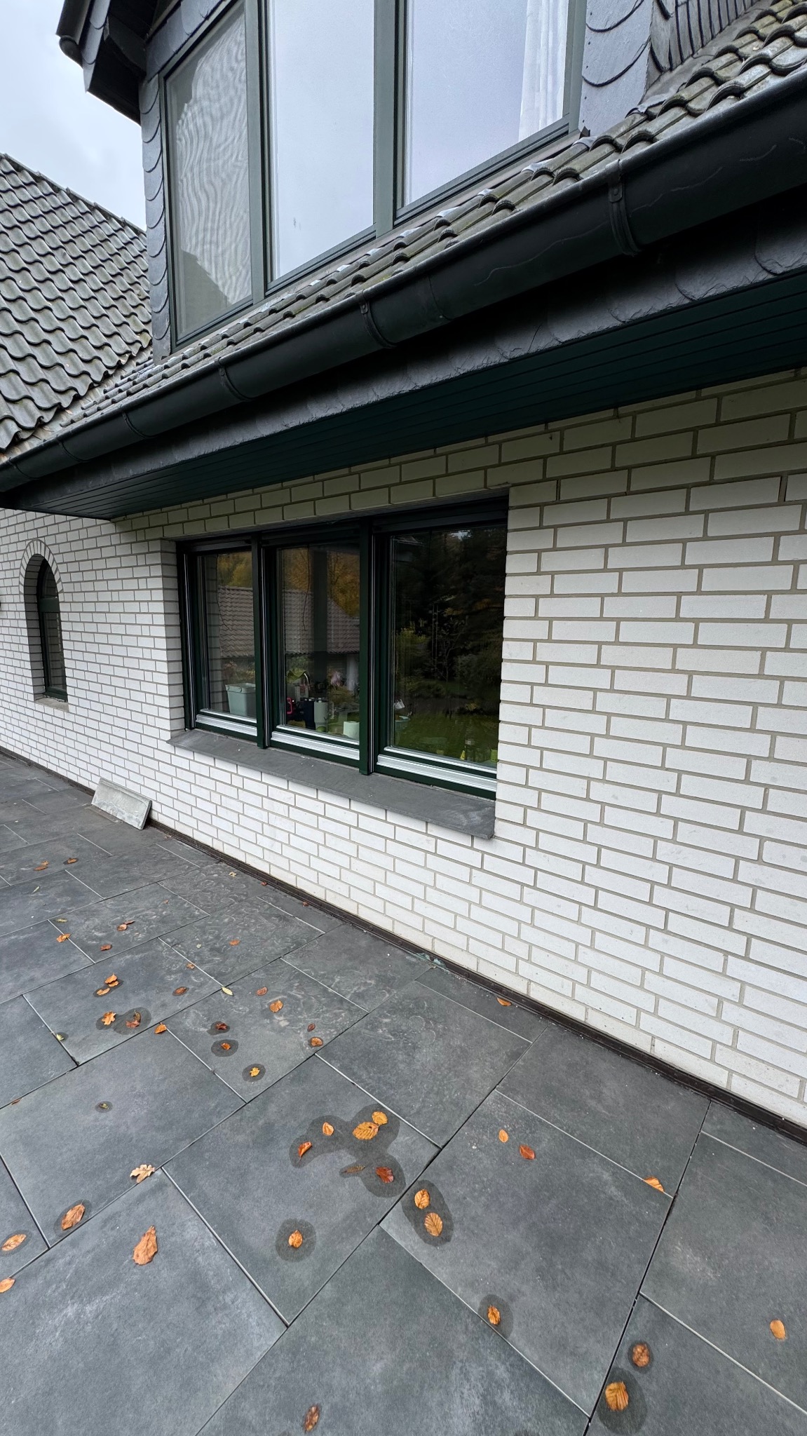 Elewacja domu z białej cegły z oknami w ciemnych ramach. Na szarym chodniku leżą opadłe liście. Widoczne okno łukowe.