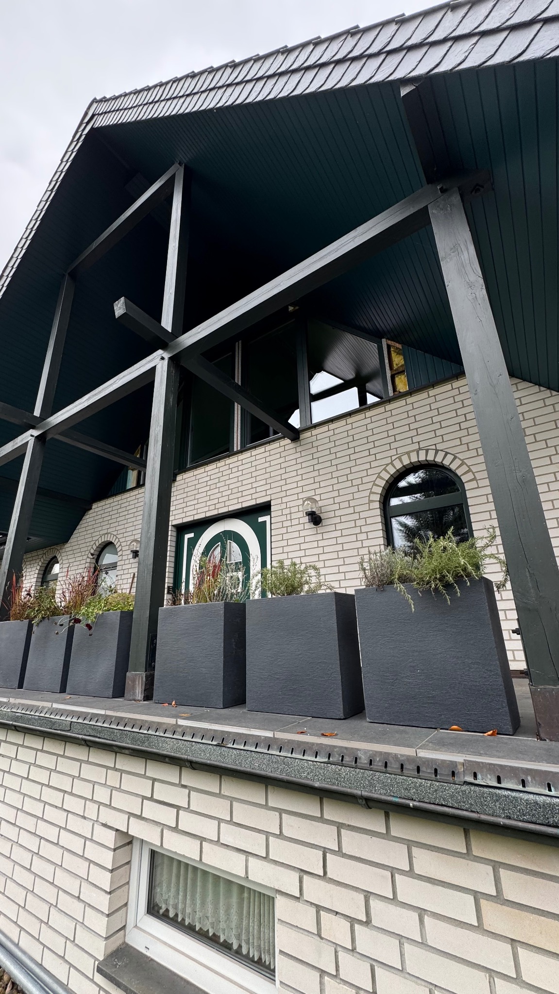 Biała fasada domu z ciemnozielonymi elementami konstrukcyjnymi i donicami. Widoczne okna z łukami i drzwi wejściowe z okrągłym oknem. Ujęcie z dołu.