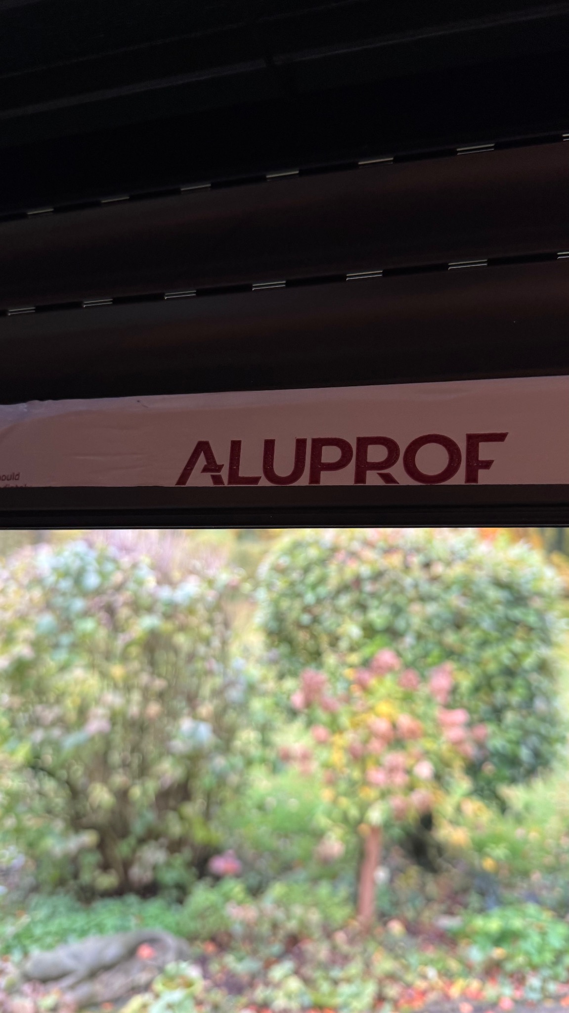 Fragment rolety zewnętrznej marki Aluprof, widok z wnętrza na zewnątrz, z rozmytym tłem zieleni. Detal produktu z bliska, z widocznym logo producenta.