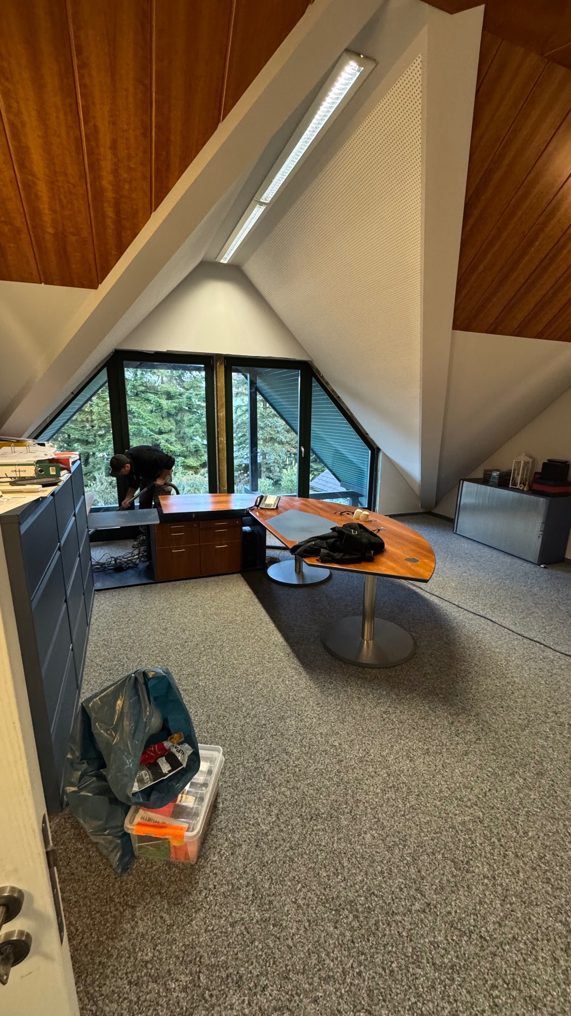 Montaż okna trapezowego w biurze na poddaszu. Widok na zieleń za oknem. Osoba montująca okno. Na podłodze torba z narzędziami.