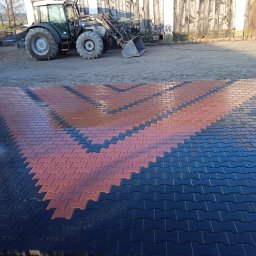 Volodymyr Koldobenko - Ułożona, wzorzysta kostka brukowa w kolorach czerwonym i czarnym na podwórku z traktorem i budynkiem gospodarczym w tle. Widoczny fragment kratki ściekowej.