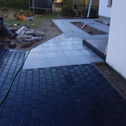 Volodymyr Koldobenko - Ułożona, czarna kostka brukowa prowadząca do domu z szarą kostką. Widoczne przygotowane podłoże i elementy ogrodu. Kąt ujęcia z dołu, podkreślający wzór kostki.