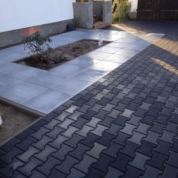 Volodymyr Koldobenko - Ułożona kostka brukowa w odcieniach szarości i grafitu, przechodząca w jasnoszare płyty chodnikowe z wyznaczonym miejscem na roślinność przy budynku.