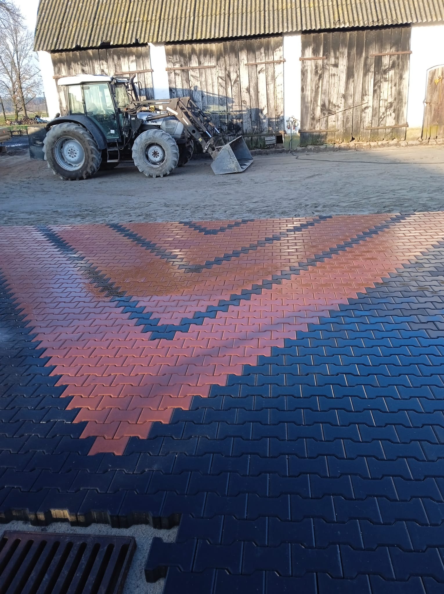 Ułożona, wzorzysta kostka brukowa w kolorach czerwonym i czarnym na podwórku z traktorem i budynkiem gospodarczym w tle. Widoczny fragment kratki ściekowej.