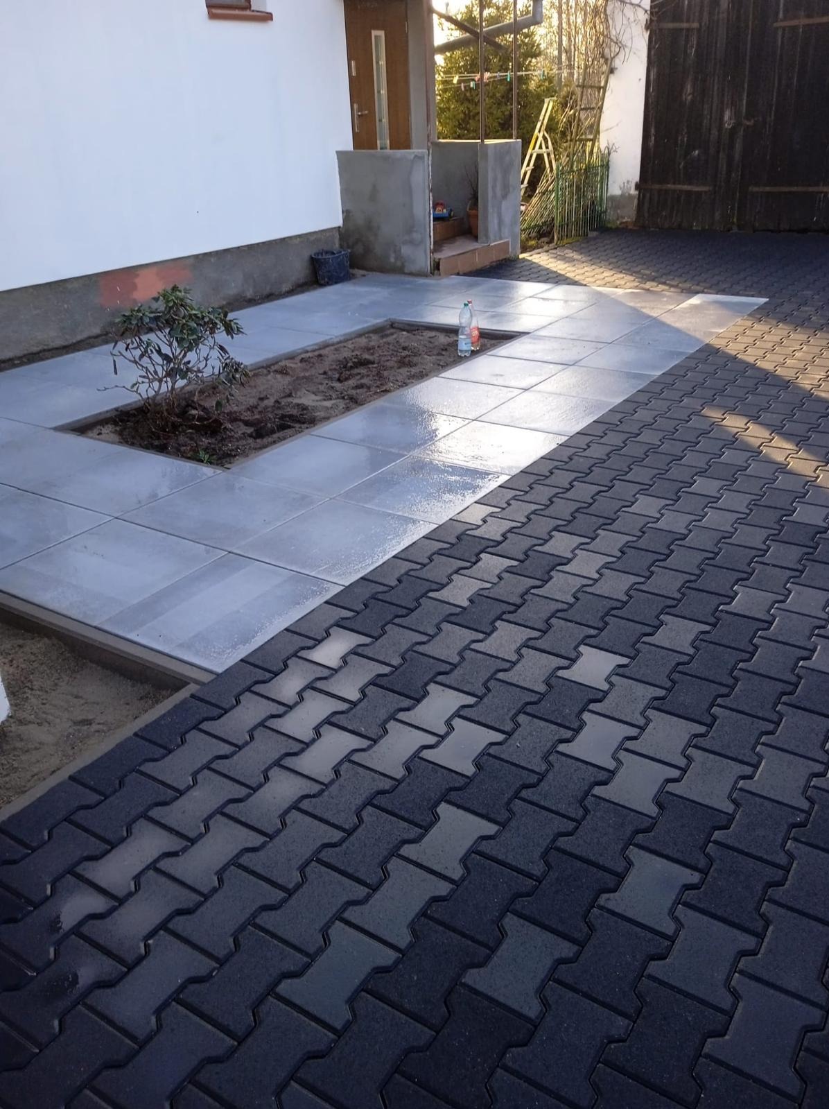 Ułożona kostka brukowa w odcieniach szarości i grafitu, przechodząca w jasnoszare płyty chodnikowe z wyznaczonym miejscem na roślinność przy budynku.