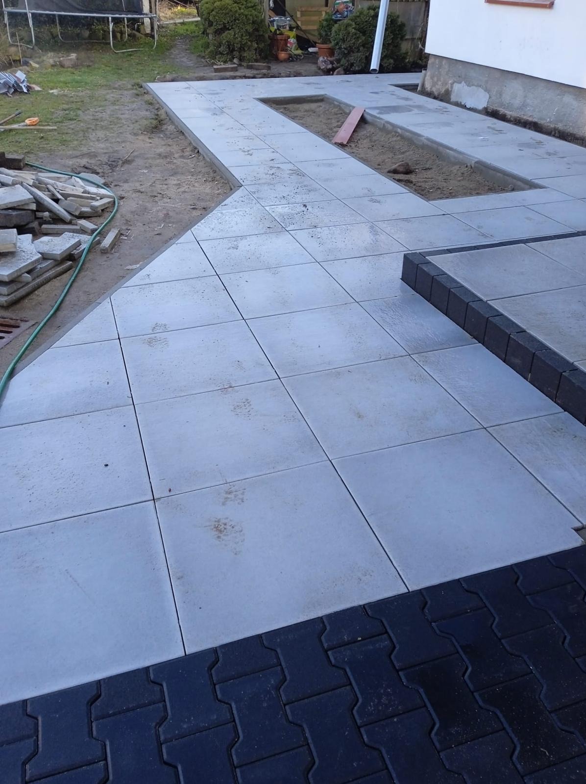 Ułożona szara kostka brukowa na posesji z czarnym obrzeżem i miejscami na rośliny. Częściowo ułożona kostka, widoczne narzędzia i materiały budowlane.