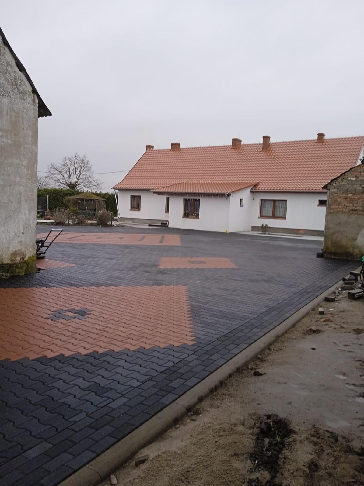 Ułożona kostka brukowa w dwóch kolorach (czarny i pomarańczowy) na podjeździe przed domem z czerwoną dachówką. Widoczny fragment budynku z prawej strony.