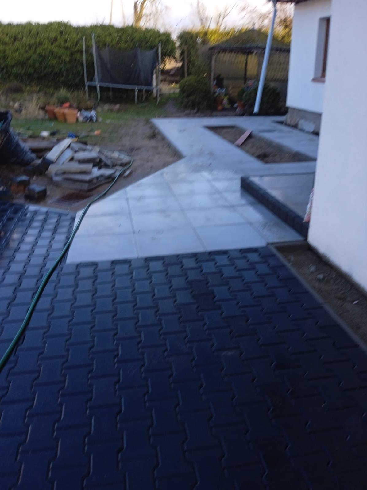 Ułożona, czarna kostka brukowa prowadząca do domu z szarą kostką. Widoczne przygotowane podłoże i narzędzia. Tło: trawnik, krzewy, trampolina.