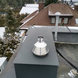 Łukasz Urbaniak - Metalowy komin wentylacyjny na płaskim dachu budynku w zimowej scenerii. Widoczne przewody i elementy instalacji. Tło stanowią ośnieżone dachy i drzewa.