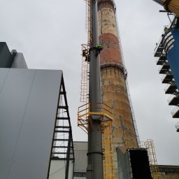 Łukasz Urbaniak - Dwóch pracowników na wysokościach, montujących nowy komin z żółtą konstrukcją, obok starego, ceglanego komina. Żuraw w tle. Szary budynek przemysłowy po lewej.