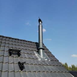 Łukasz Urbaniak - Szary komin wentylacyjny na dachu z dachówki, z zamontowanymi stopniami kominiarskimi, na tle błękitnego nieba. Widok z dołu, podkreślający wysokość komina.