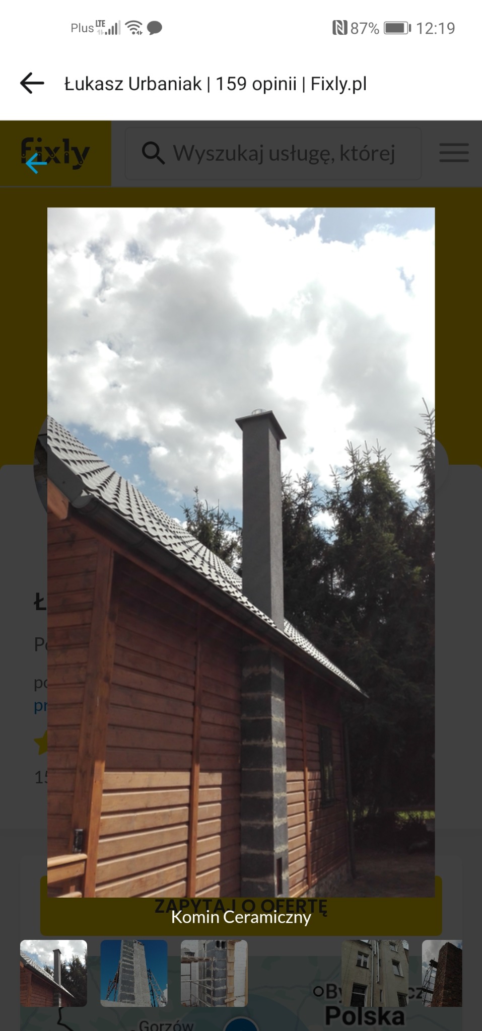 Komin ceramiczny przy drewnianym domu z szarą dachówką, widok z dołu na tle błękitnego nieba z chmurami. Detal konstrukcji kominowej.