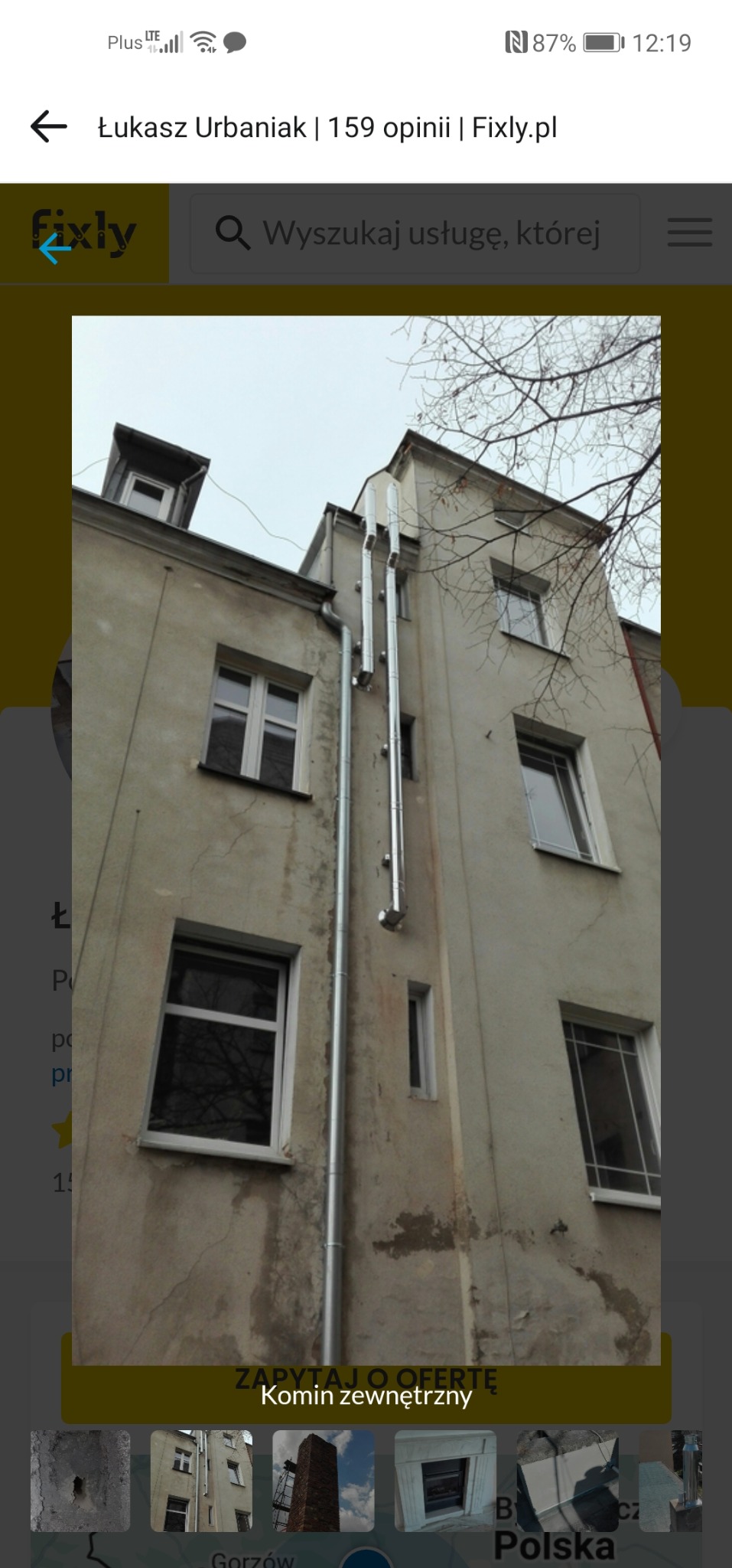 Szarego budynku z oknami i zewnętrznym, metalowym kominem. Widok z dołu, z fragmentem drzewa w tle. Komin biegnie wzdłuż ściany budynku.