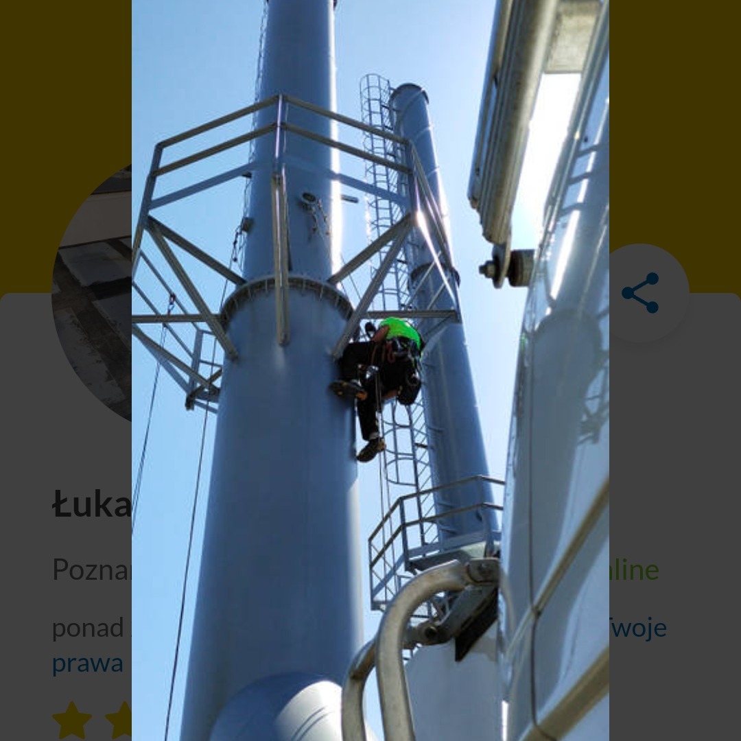 Pracownik w uprzęży, na wysokości, podczas inspekcji komina przemysłowego z metalowymi drabinkami i platformami, na tle błękitnego nieba. Widok z dołu.