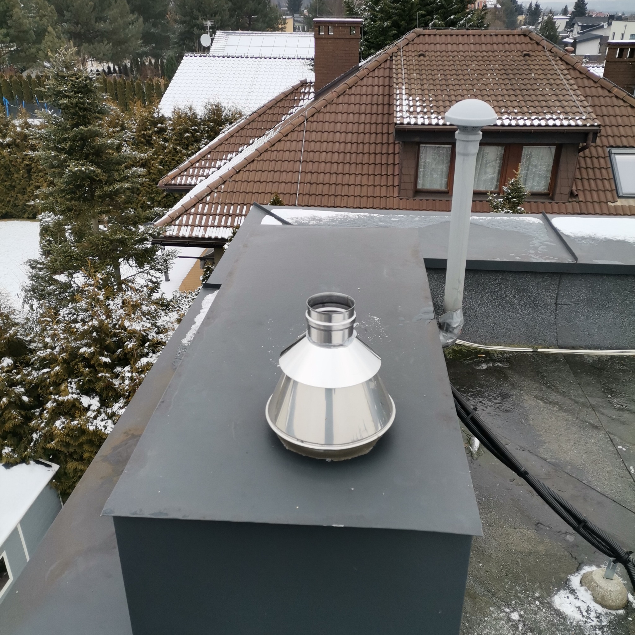 Metalowy komin wentylacyjny na płaskim dachu budynku w zimowej scenerii. Widoczne przewody i elementy instalacji. Tło stanowią ośnieżone dachy i drzewa.