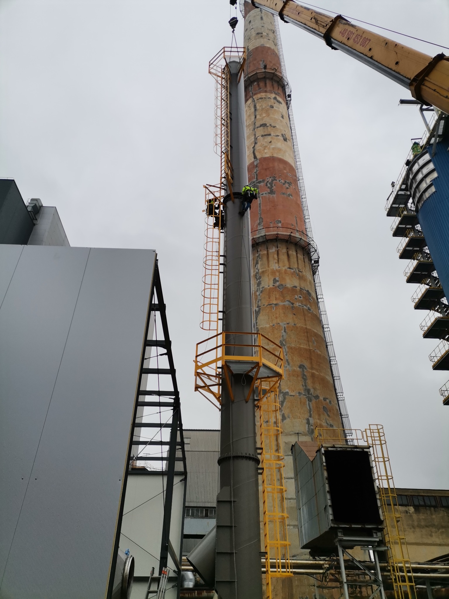 Dwóch pracowników na wysokościach, montujących nowy komin z żółtą konstrukcją, obok starego, ceglanego komina. Żuraw w tle. Szary budynek przemysłowy po lewej.