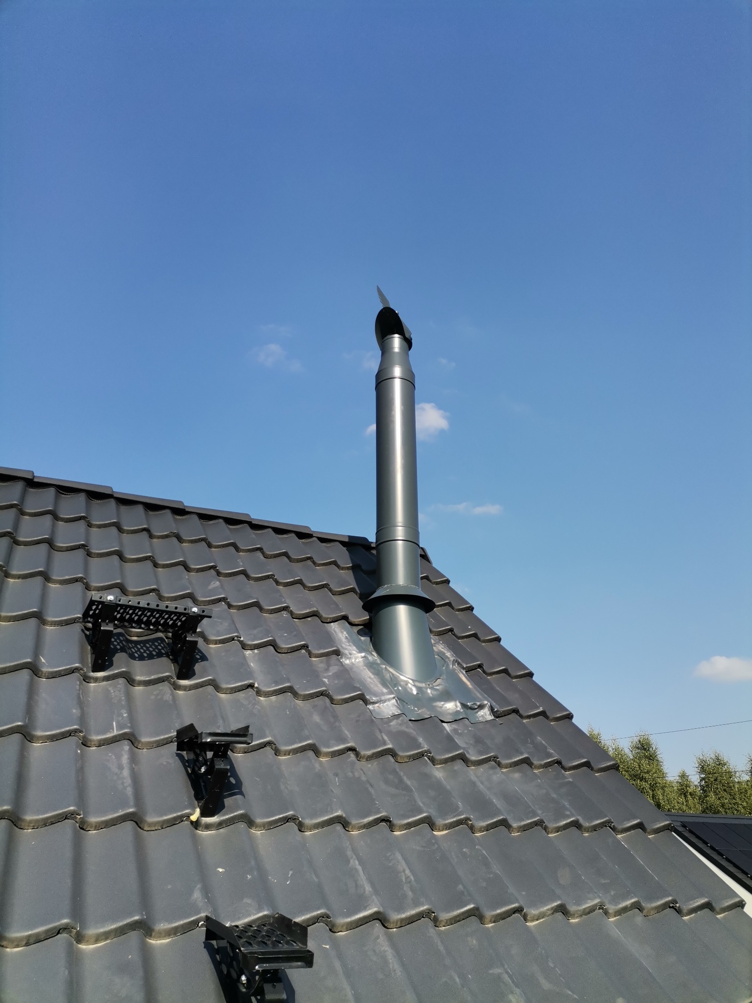 Szary komin wentylacyjny na dachu z dachówki, z zamontowanymi stopniami kominiarskimi, na tle błękitnego nieba. Widok z dołu, podkreślający wysokość komina.
