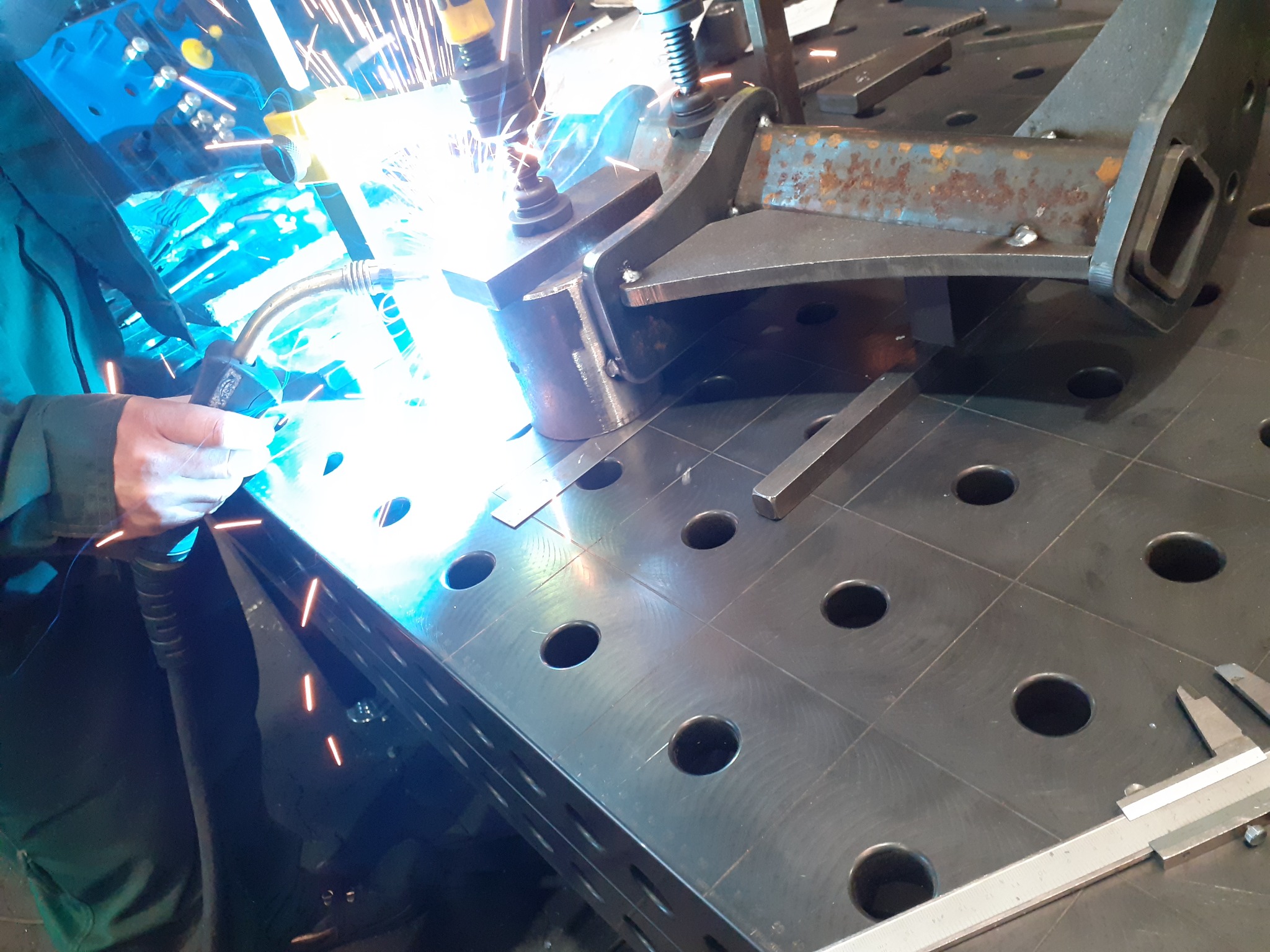 Spawanie elementu metalowego na perforowanym stole spawalniczym. Widoczne iskry i narzędzia pomiarowe. Praca w toku, zbliżenie na proces łączenia metali.