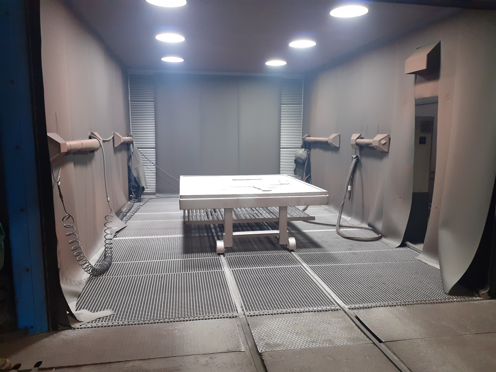 Biały metalowy stół na kółkach wewnątrz lakierni proszkowej z kratą podłogową i oświetleniem sufitowym. Widoczne dysze natryskowe na ścianach.