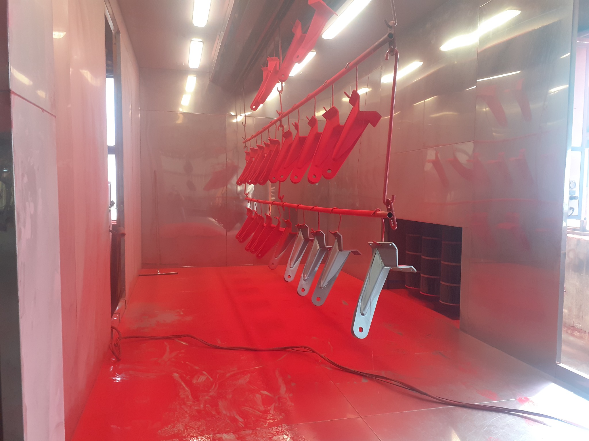 Metalowe elementy konstrukcyjne zawieszone w kabinie lakierniczej, część pomalowana na czerwono, część w kolorze metalu. Widoczne filtry i oświetlenie.