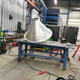 Weldingteam - Metalowy stożek na stole warsztatowym w hali produkcyjnej, widoczny sprzęt spawalniczy i suwnica. Element konstrukcyjny w trakcie obróbki.