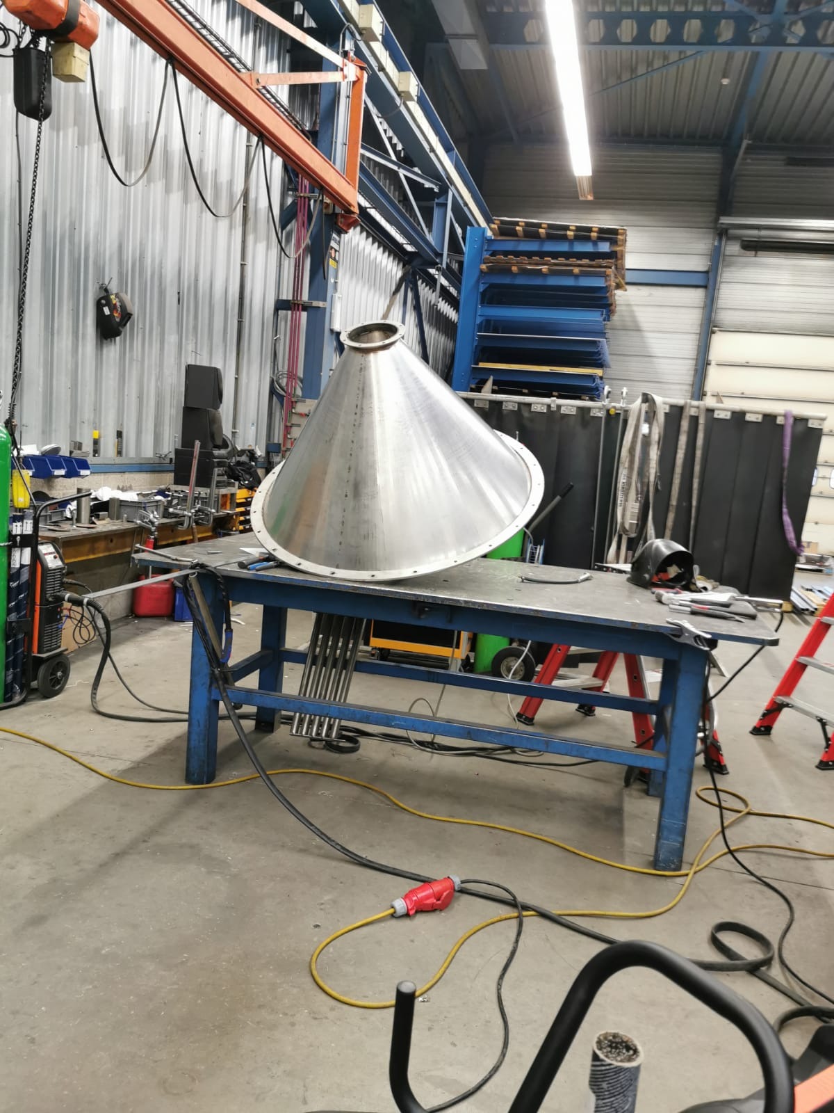 Metalowy stożek na stole warsztatowym w hali produkcyjnej, widoczny sprzęt spawalniczy i suwnica. Element konstrukcyjny w trakcie obróbki.