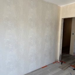 Vitalii Usenko - Ściana po malowaniu z delikatnym wzorem, widoczne zabezpieczone podłogi i fragment otworu drzwiowego w trakcie prac wykończeniowych.