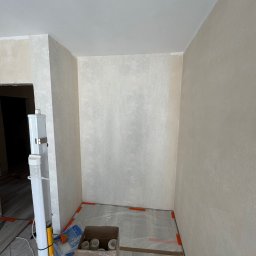 Vitalii Usenko - Świeżo pomalowane, beżowe ściany w rogu pokoju, zabezpieczone folią malarską. Widoczny karton z akcesoriami malarskimi i poziomica. Podłoga częściowo wyłożona panelami.