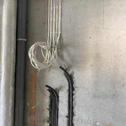 FixHouse Grzegorz Rzepa - Surowa ściana z instalacją elektryczną: białe kable spięte u góry, czarne rury karbowane w bruzdach na dole. Obok rury kanalizacyjne. Etap prac wykończeniowych.