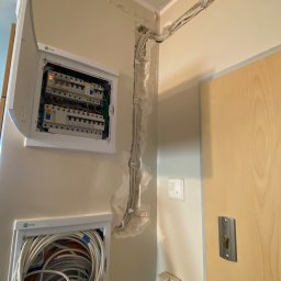 FixHouse Grzegorz Rzepa - Rozprowadzenie instalacji elektrycznej w ścianie, widoczne kable w korytkach i skrzynka rozdzielcza z bezpiecznikami. Prace instalacyjne wewnątrz budynku.