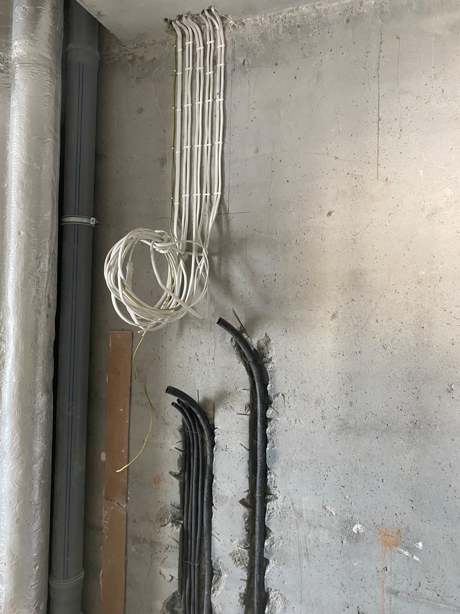 Surowa ściana z instalacją elektryczną: białe kable spięte u góry, czarne rury karbowane w bruzdach na dole. Obok rury kanalizacyjne. Etap prac wykończeniowych.
