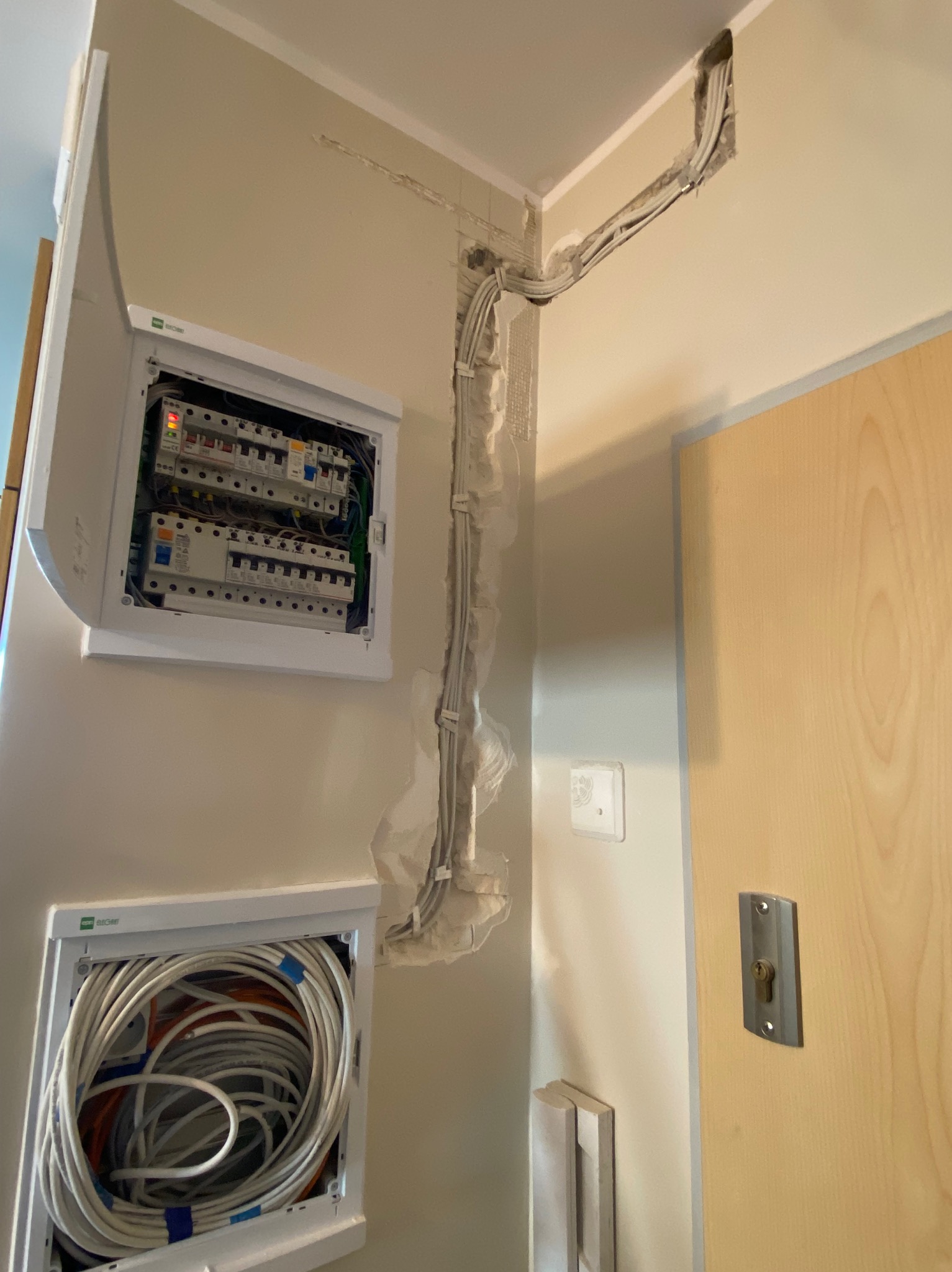 Rozprowadzenie instalacji elektrycznej w ścianie, widoczne kable w korytkach i skrzynka rozdzielcza z bezpiecznikami. Prace instalacyjne wewnątrz budynku.