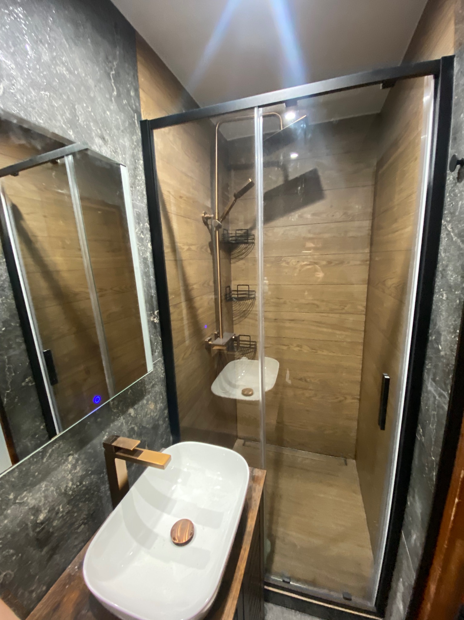 Nowoczesna łazienka z kabiną prysznicową typu walk-in, drewnianymi panelami ściennymi i umywalką nablatową z miedzianą armaturą. Lustro z podświetleniem.