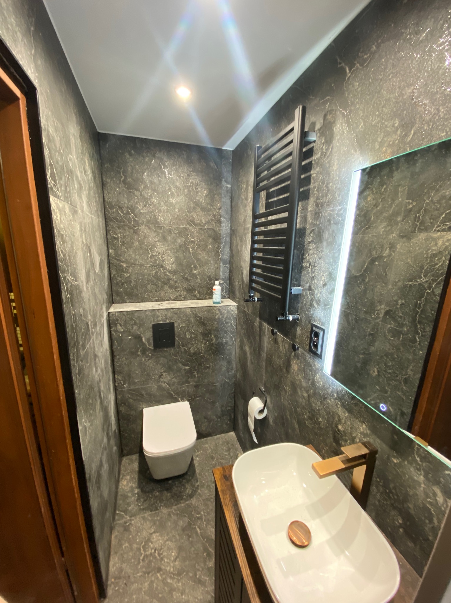 Nowoczesna łazienka z szarymi płytkami imitującymi kamień, podświetlanym lustrem i drewnianą szafką pod umywalką. Minimalistyczny design z grzejnikiem drabinkowym.