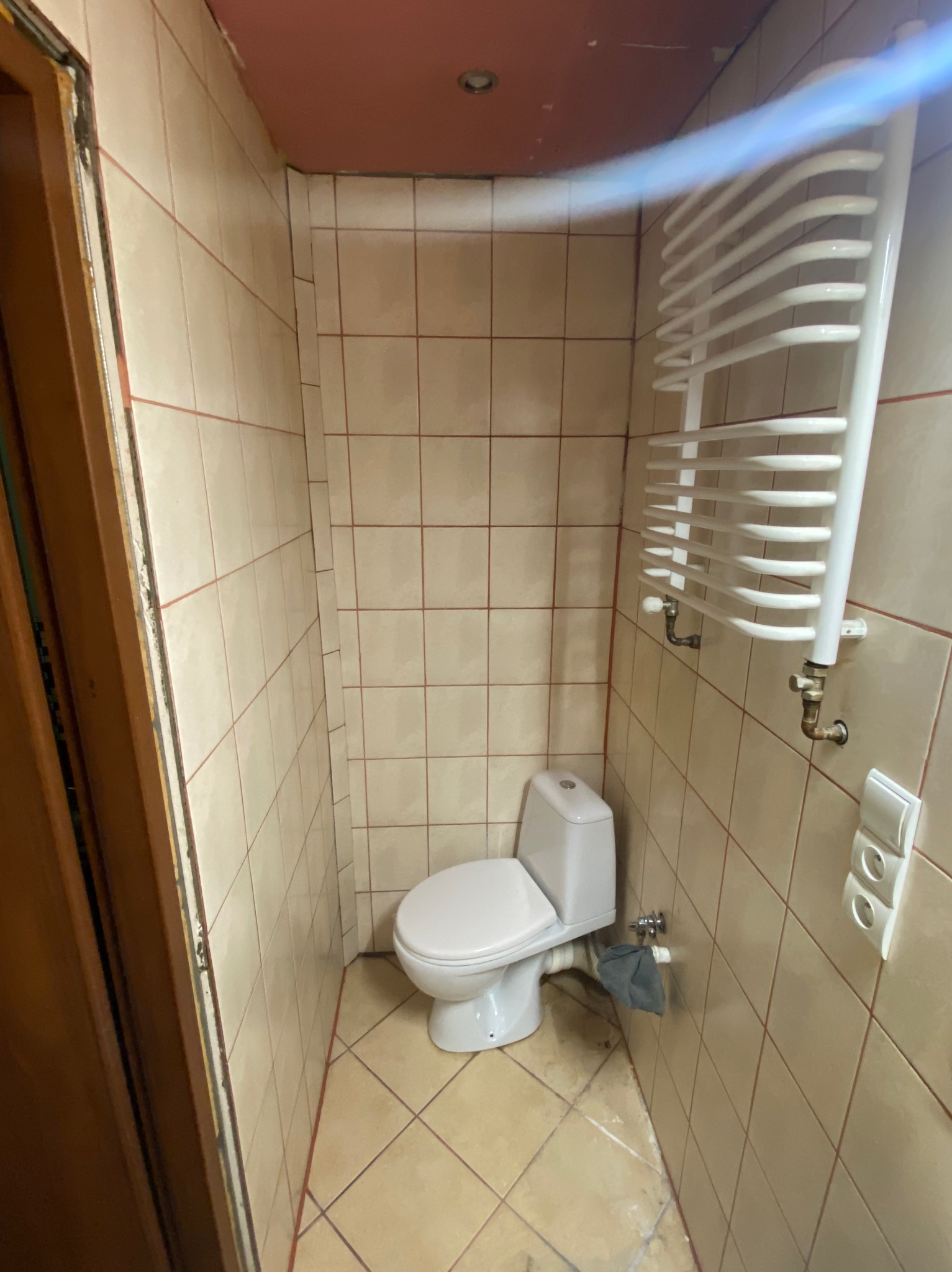 Wykończona łazienka z beżowymi płytkami, białą toaletą i grzejnikiem drabinkowym. Widoczne detale: brązowe obramowanie drzwi, różowy sufit i potrójny włącznik światła.