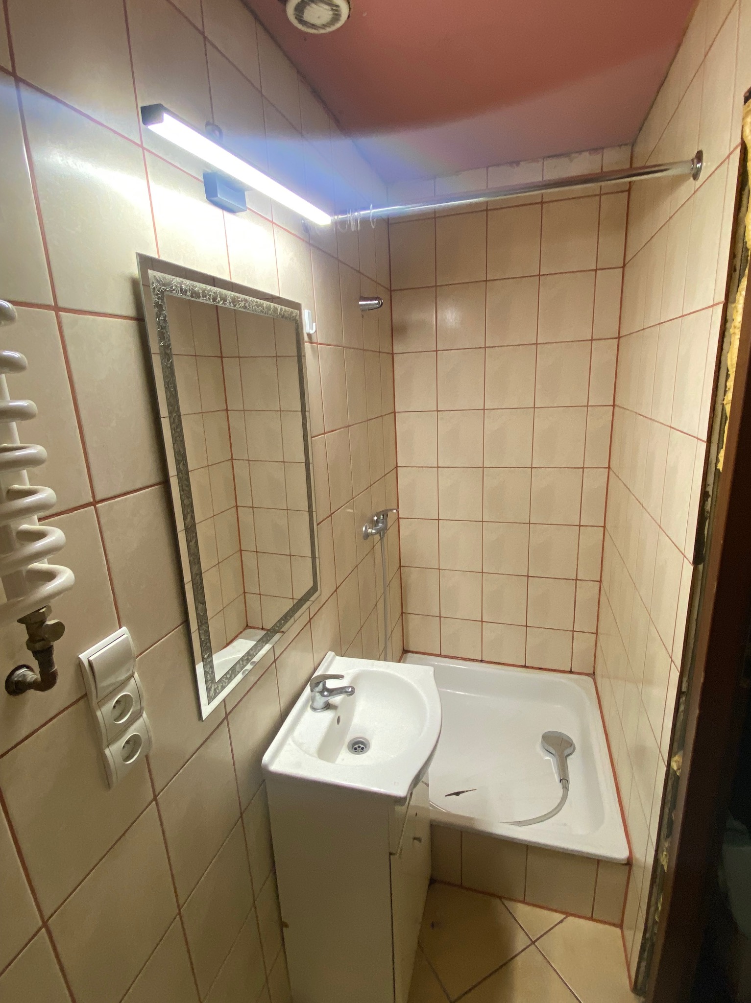 Jasna łazienka z beżowymi płytkami, kabiną prysznicową i umywalką na postumencie. Widoczne lustro w ozdobnej ramie i grzejnik drabinkowy.