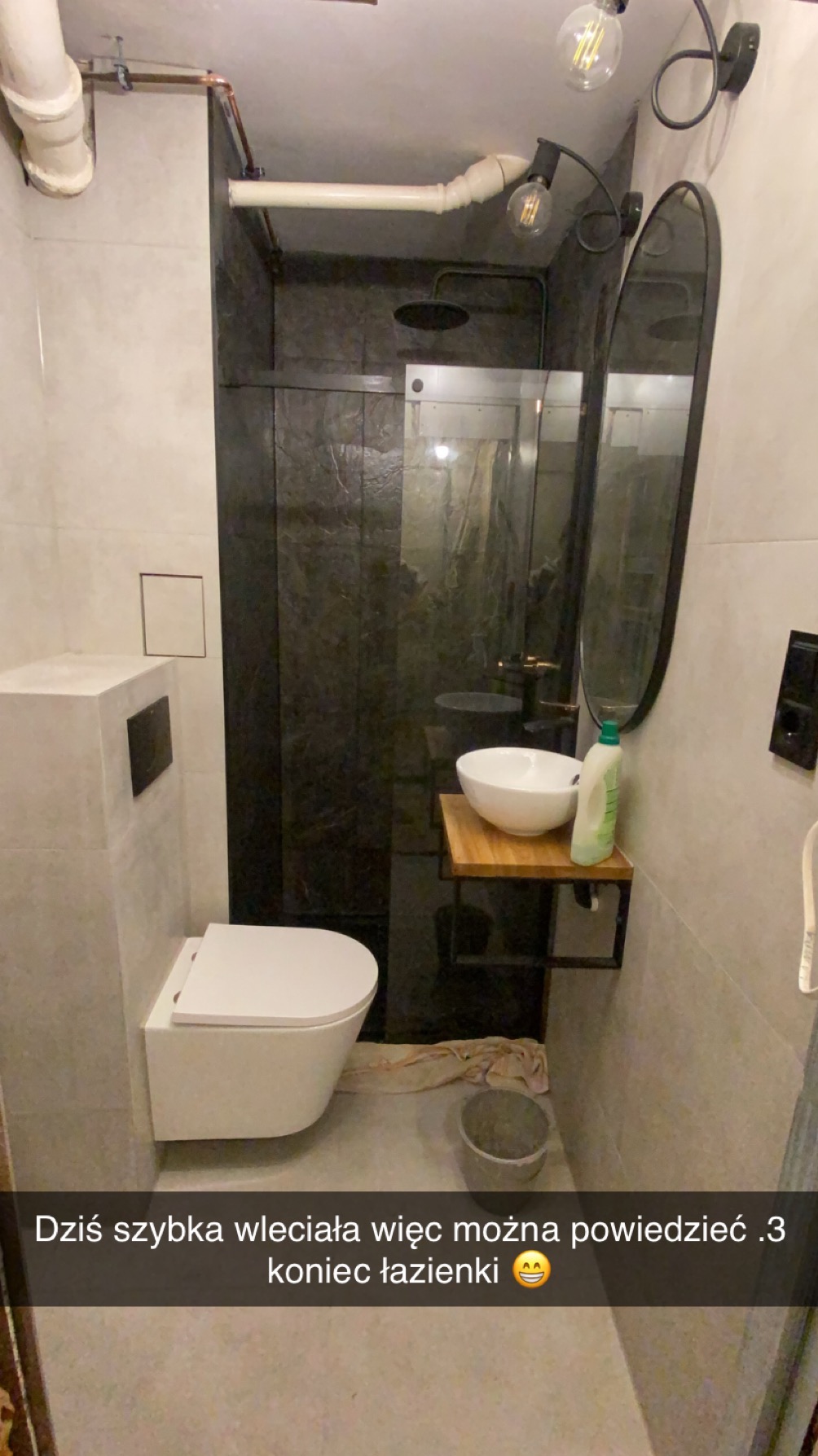 Wykończona łazienka w szarościach z kabiną prysznicową typu walk-in, umywalką na drewnianym blacie i podwieszaną toaletą. Widoczne rury pod sufitem i designerskie oświetlenie.