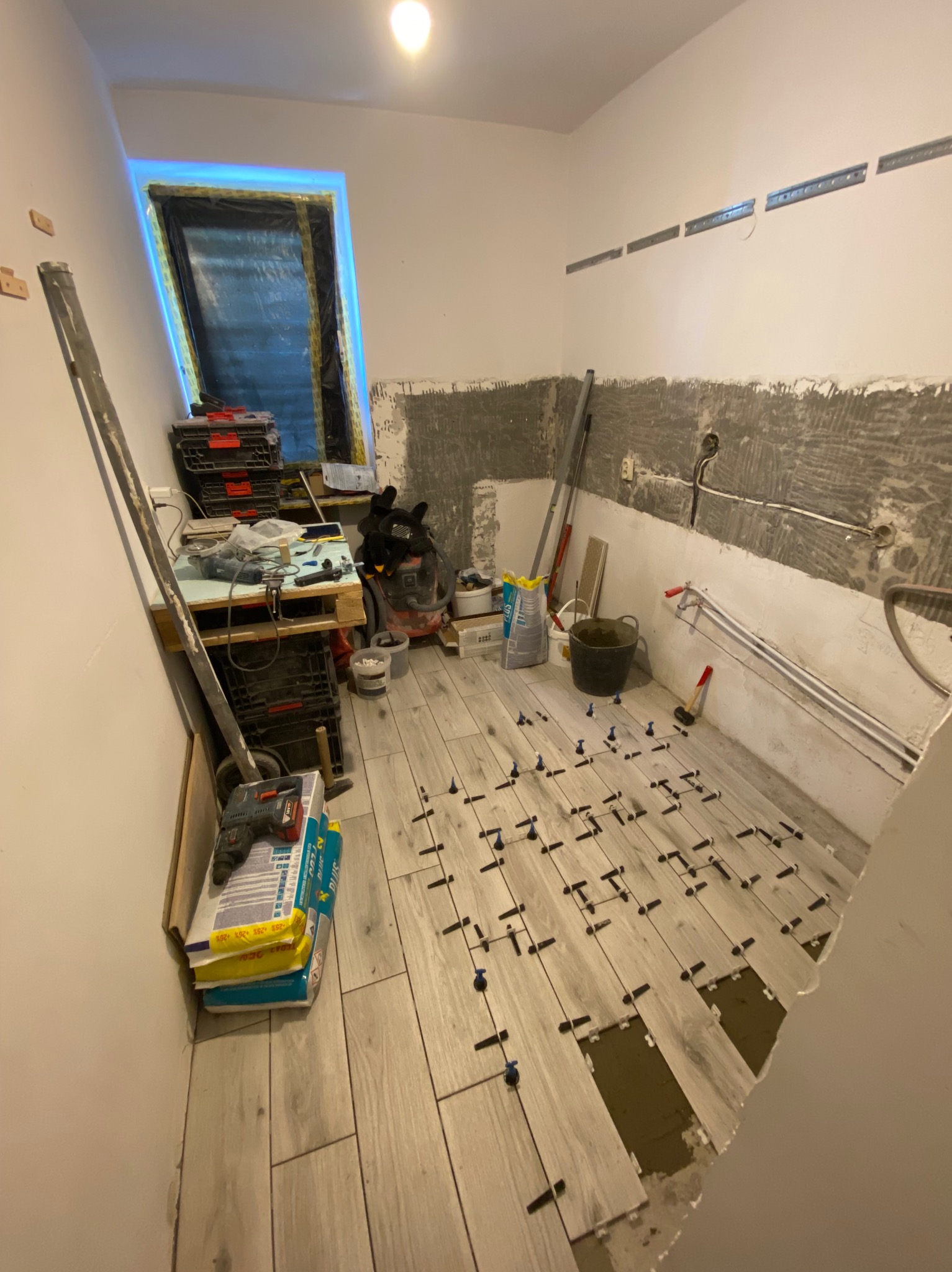 Układanie płytek podłogowych w łazience, widoczne krzyżyki dystansowe, narzędzia i materiały budowlane. Częściowo wykończona ściana z widocznymi rurami.