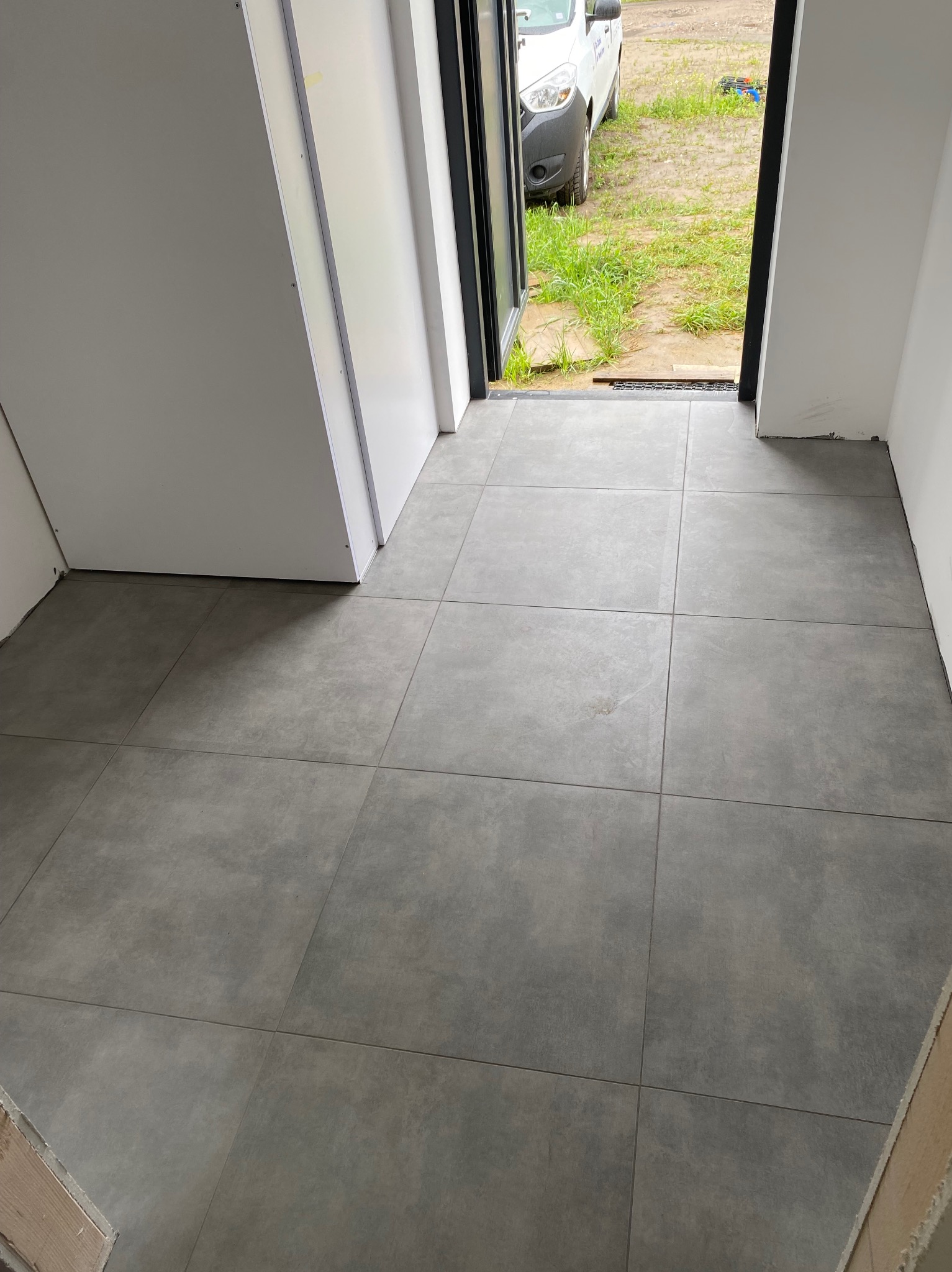 Ułożone, szare płytki podłogowe w korytarzu, widok z góry. Na zewnątrz widoczny trawnik i zaparkowany samochód dostawczy. Minimalistyczne wykończenie.