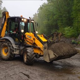 JACEK WANTUŁA Firma Handlowo-Usługowa JAC-KOP - Żółta koparko-ładowarka JCB 3CX z pełną łyżką ziemi na błotnistym terenie, w tle hałda żwiru i ściana z betonowych prefabrykatów, otoczona zielenią.